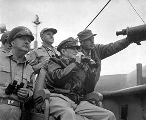 macarthur is seated, wearing his field marshal\'s hat and a bomber jacket, and holding a pair of binoculars. four other men also carrying binoculars stand behind him.