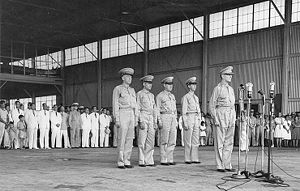 macarthur stands in uniform at four microphones on stands. behind him four men in army uniforms stand at attention. there are viewed by a large crowd of well-dressed men, women and children in skirts, suits and uniforms.