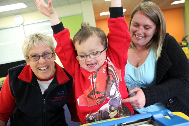 preschool boy holds up his hands in excitement after using the mountbatten brailler. his teacher sits on his right, and mother on his left.