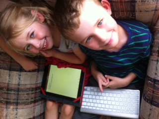 young boy and girl sitting. the girl has the ipad on her knee, and the boy has a wireless keyboard.