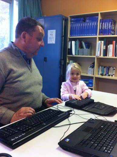 young girl standing next to a man sitting at a desk. the young girl positions her hands on a refreshable braille display.