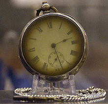 photograph of a brass pocket watch on a stand, with a silver chain curled around the base. the watch\'s hands read 2:28.