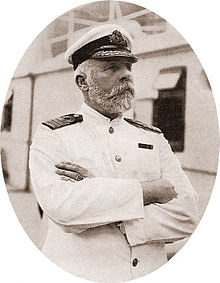 photograph of a bearded man wearing a white captain\'s uniform, standing on a ship with his arms crossed.