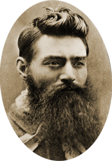 head of a young man with a long, untrimmed beard, and with hair cropped above the ears, but longer and slicked up and back on the top. his moustache and beard are so long that his mouth and shirt front can barely be seen. his eyes look over the viewer\'s right shoulder.