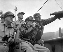 macarthur is seated, wearing his field marshal\'s hat and a bomber jacket, and holding a pair of binoculars. four other men also carrying binoculars stand behind him.