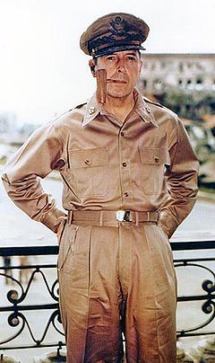 macarthur in khaki trousers and open necked shirt with five-star-rank badges on the collar. he is wearing his field marshal\'s cap and smoking a corncob pipe.